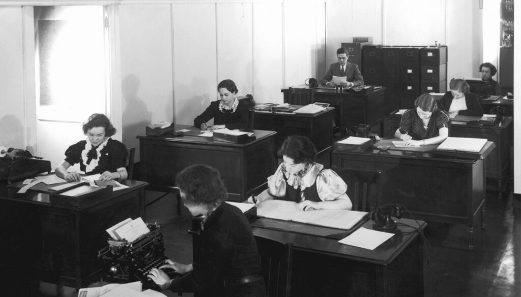 1950s black and white photo of office workers sitting and writing at their desks