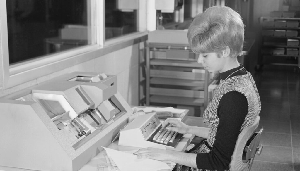 1950s black and white photo of young woman working on a typewriter in front of lab screens