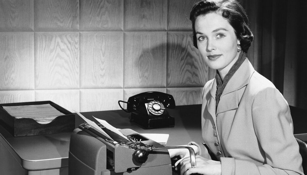 1950s black and white photo of a woman sitting at a typewriter looking at the camera