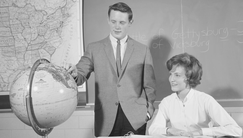 1950s black and white photo of a student pointing at a globe next to a smiling teacher
