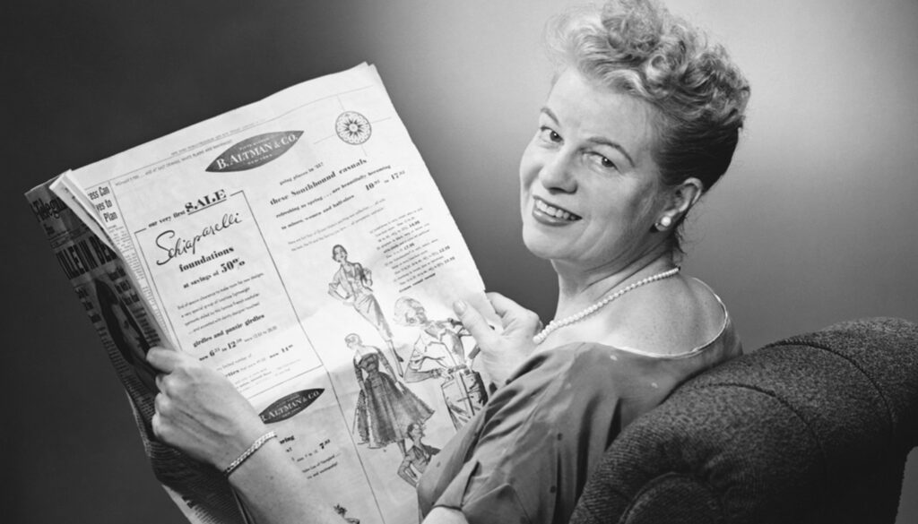 1950s black and white photo of woman sitting in a recliner smiling while looking at a newspaper