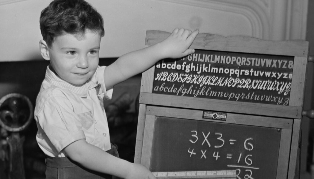 1950s black and white photo of a young boy showing off his math skills on a chalkboard