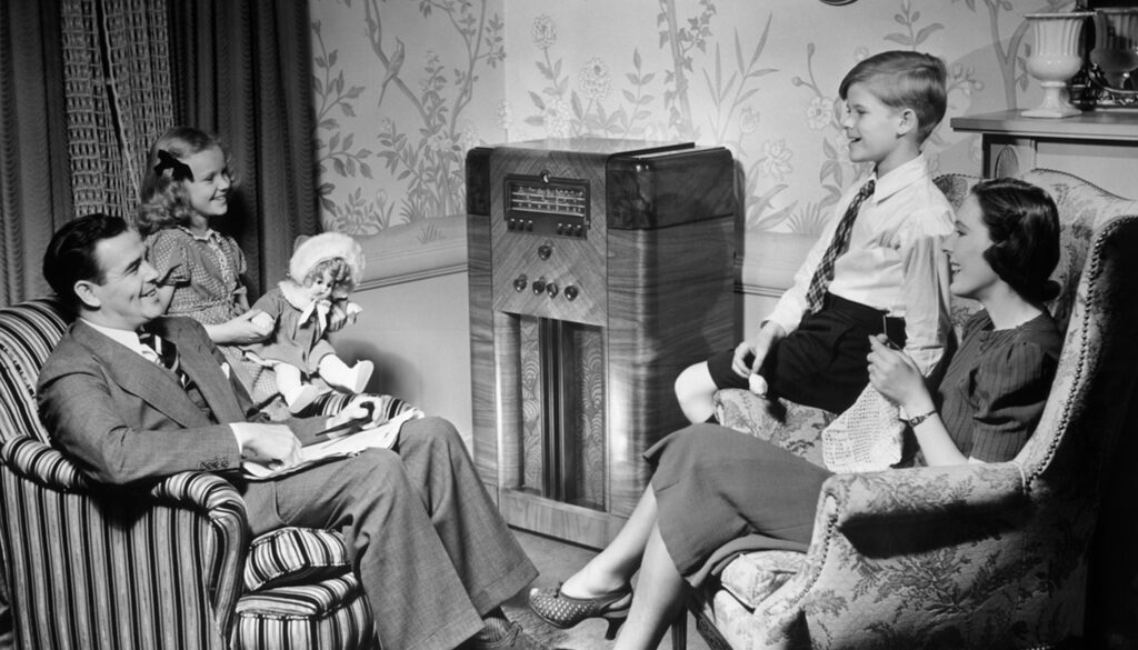 1950s black and white photo of a family with a dad, mom, daughter, and son gathered around a radio